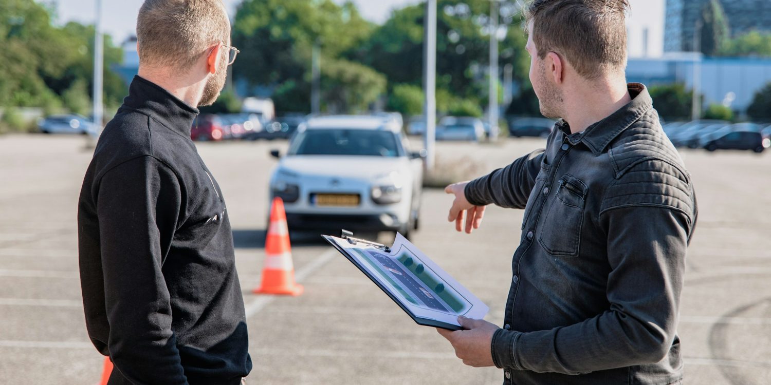 Inštruktor autoškoly trénuje študenta na parkovisku s kužeľmi a bielym autom počas hodiny autoškoly